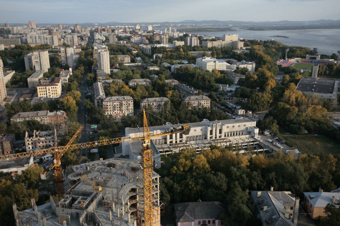 Aerial view of Khabarovsk showing Soviet-era apartment blocks, new construction cranes, and the Amur River in the distance.