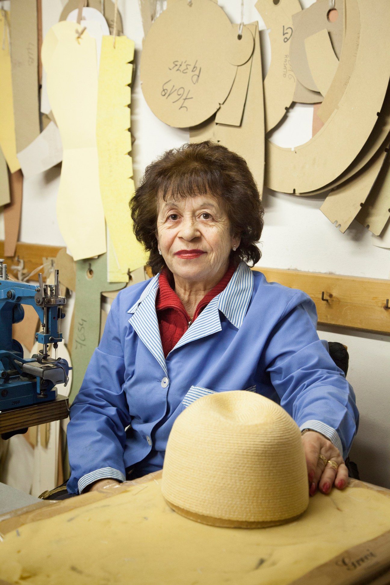 Veteran seamstress Maria D'Areglia sits at her workstation with wooden hat blocks and a straw hat.