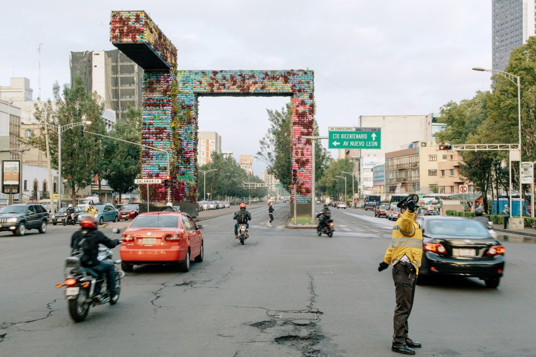 Large vertical eco-sculpture with colorful flowers and plants frames a Mexico City street with traffic and motorcyclists.