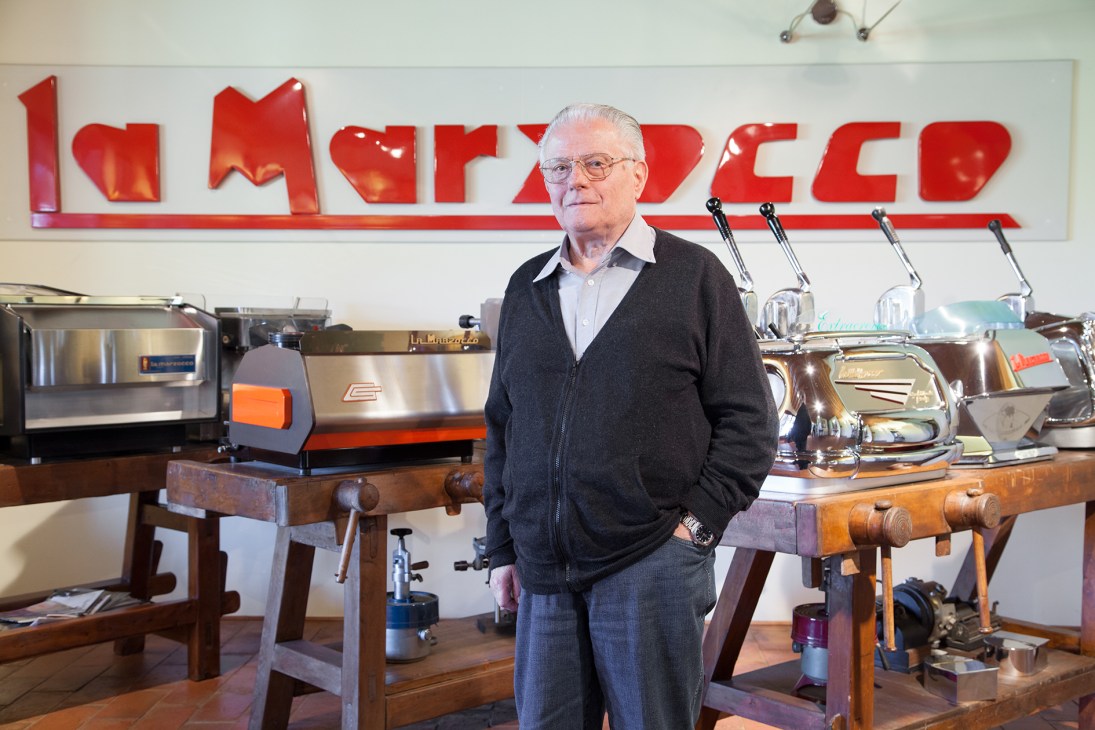 Piero Bambi, 81-year-old honorary president of La Marzocco, stands in front of espresso machines with the company's red logo visible behind him.