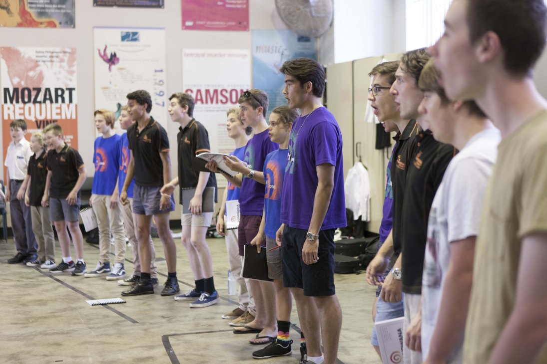 Young choir members wearing purple and black uniforms stand in a line during rehearsal in an indoor space with posters visible on the walls.