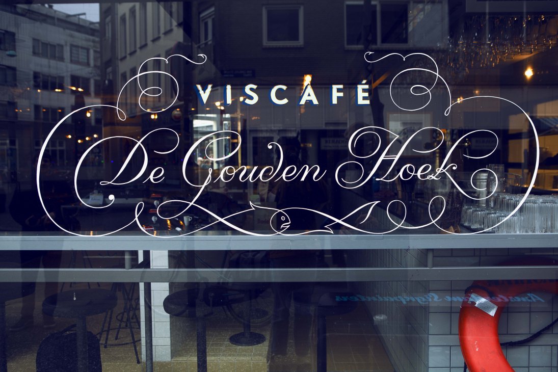 Illuminated neon cursive lettering reading "V.I.S CAFÉ De Gouden Hoek" on a cafe window at night