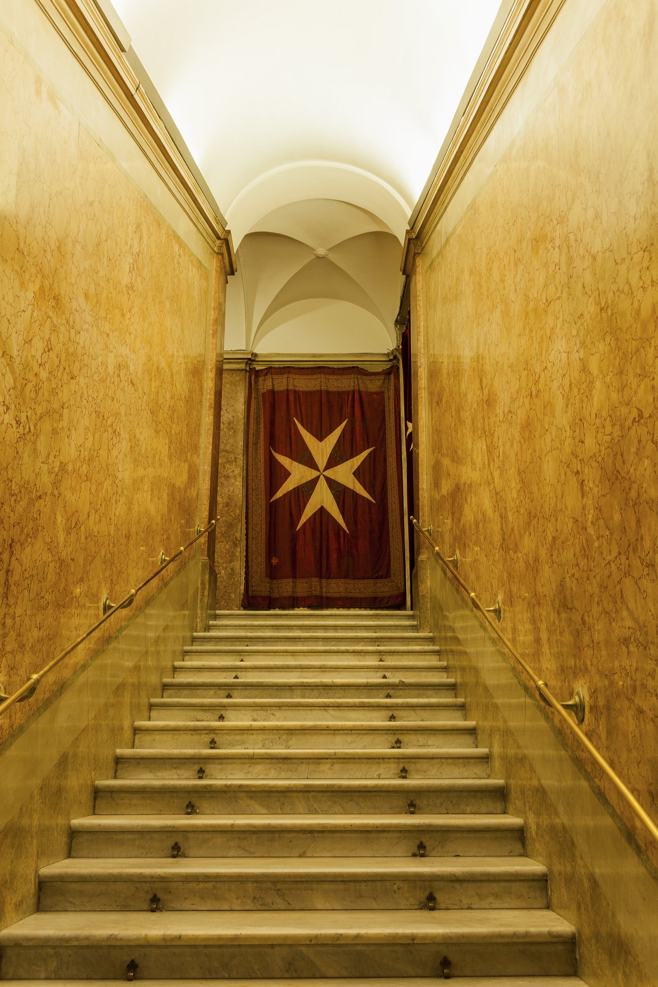 Stone staircase with handrails leading up to a red door with a gold Maltese cross in a yellow-walled corridor.