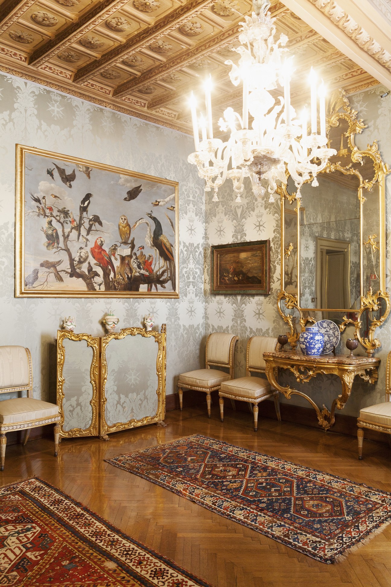Ornate reception room with gilded rococo furniture, white chandelier, patterned wallpaper, and classical paintings at Palazzo Magistrale.