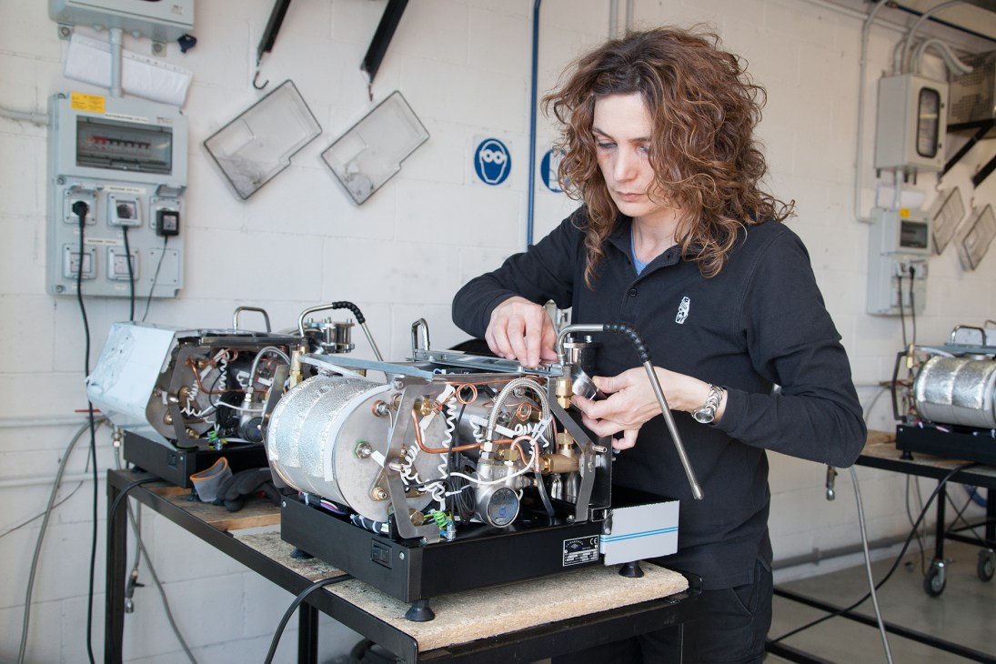A technician assembles internal components of a La Marzocco GS/3 espresso machine in a factory workshop.