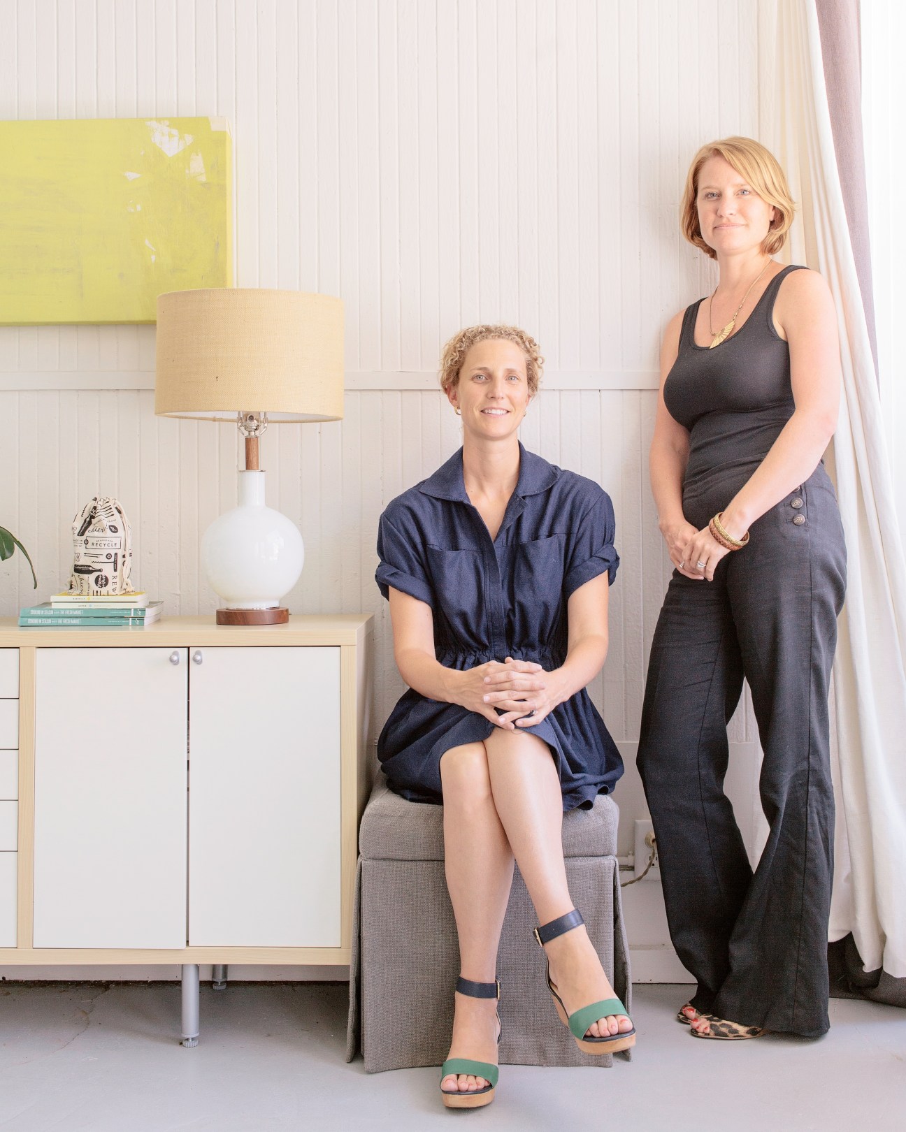 Two women posing in a bright studio space with modern furniture and a yellow artwork on the wall.