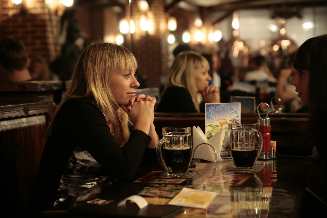 Woman with blonde hair sitting at a bar counter with a drink, looking thoughtful in a dimly lit venue.