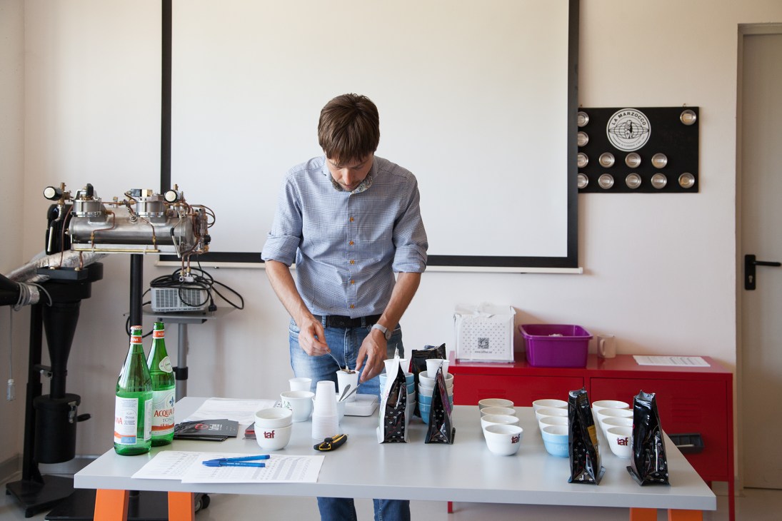 Man in blue shirt conducting coffee tasting at a lab table with espresso machine and samples.