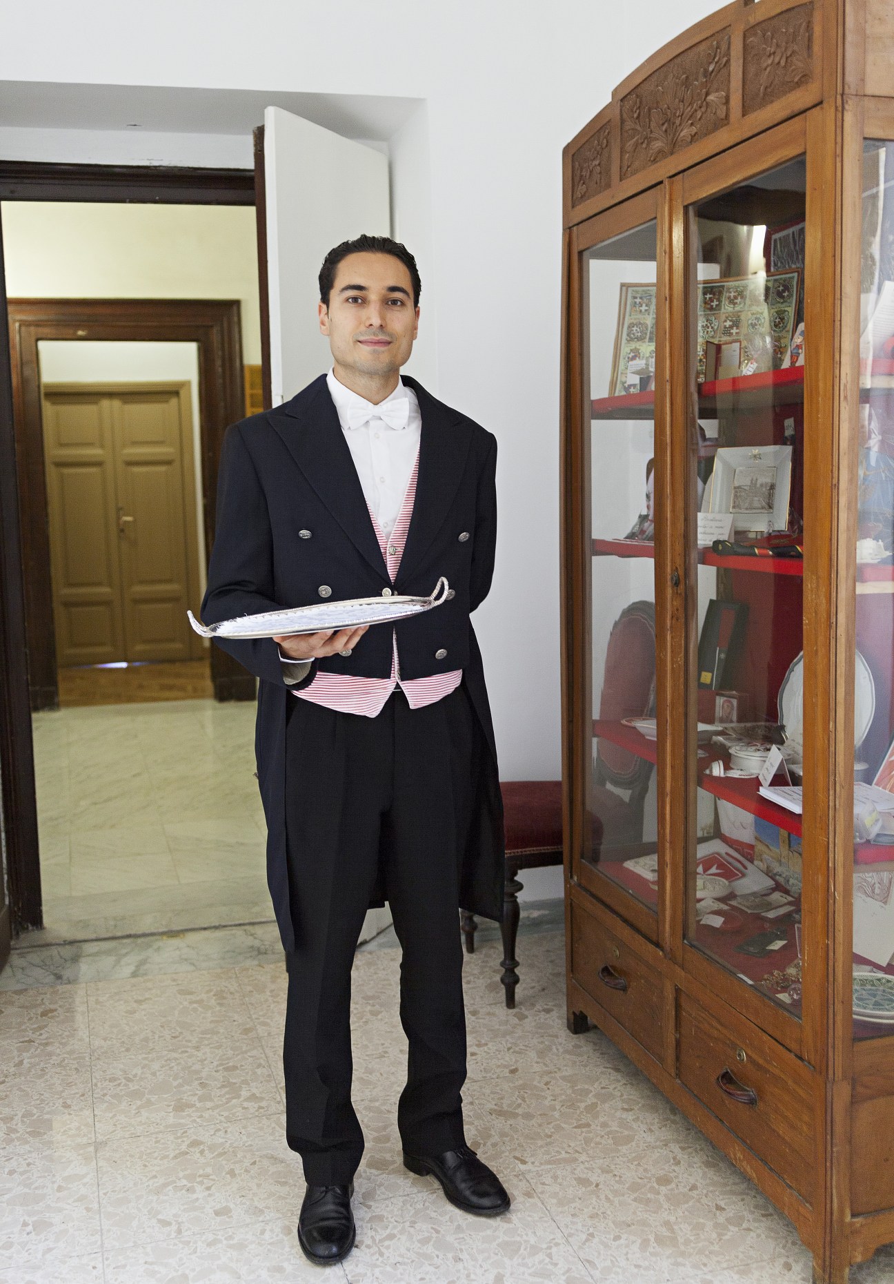 Cameriere Gabriele Pugliese in formal black tailcoat with white shirt and red striped vest, holding a silver platter in a palace corridor.