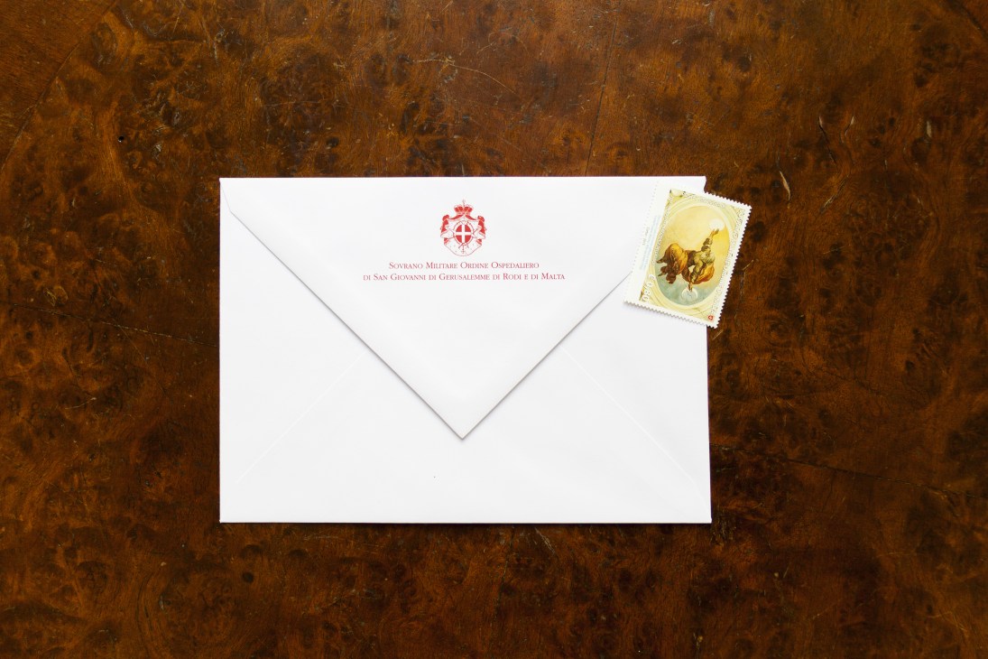 An official envelope from the Sovereign Military Order of Malta with its red emblem, displayed with a vintage stamp on a wooden surface.