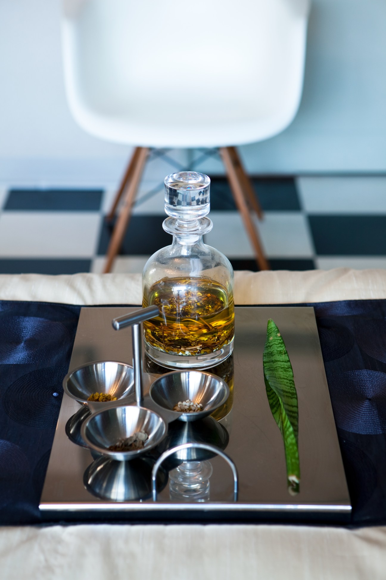 Glass bottle of amber liquid with small metal bowls on a tray with green leaf detail and white chair blurred in background.