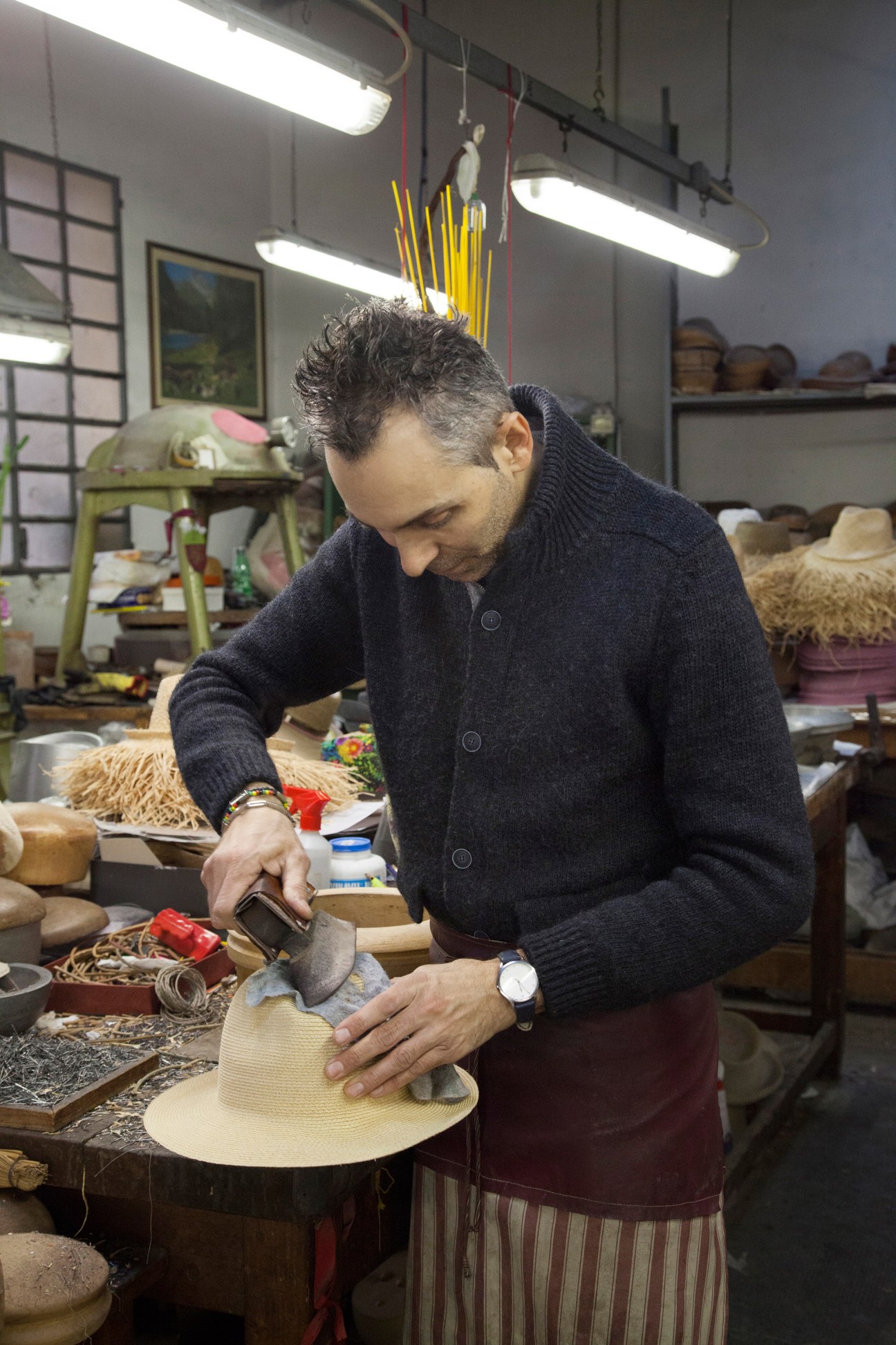 A craftsman irons the crown of a straw hat in a workshop filled with hat-making materials and tools.