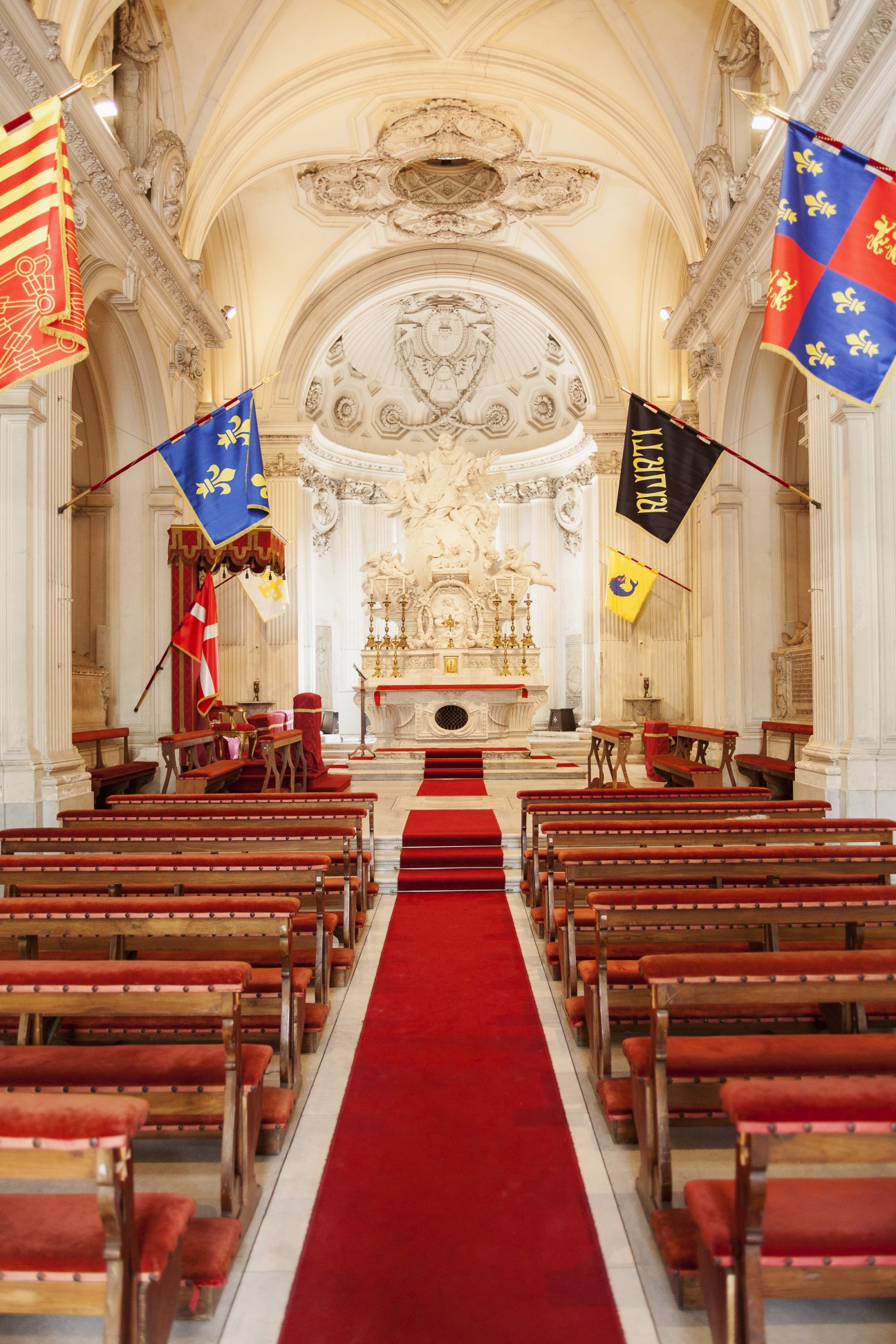 Interior of a baroque chapel with ornate ceiling, red carpet, wooden pews, and flags hung from columns.
