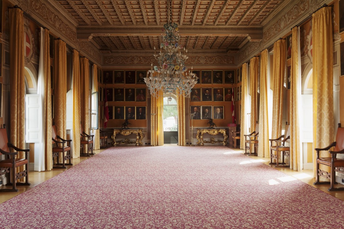 Ornate baroque hall with coffered ceiling, crystal chandelier, gilt furniture, and portraits lining the walls.
