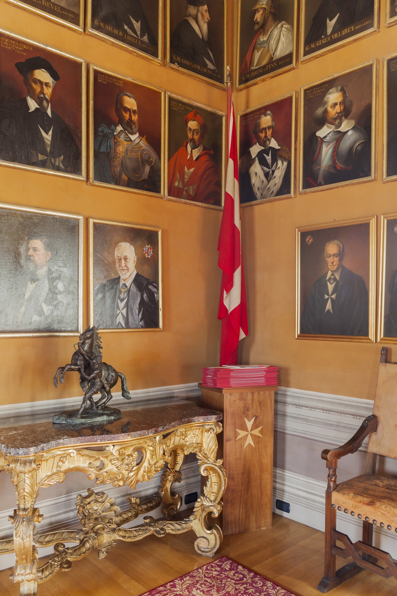 Gallery of framed portraits of grand masters hanging on ochre walls with ornate frames, a red and white flag, gilded furniture, and a bronze horse sculpture.
