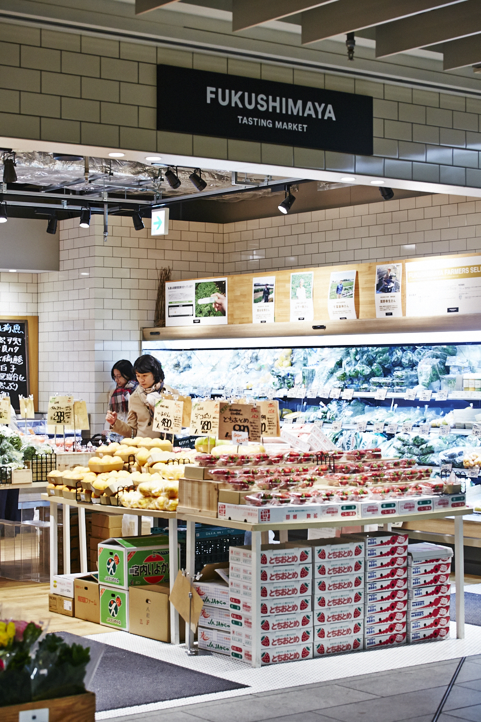 Fukushimaya Tasting Market produce counter displaying fresh fruits and vegetables with producer profile photos on the wall above.