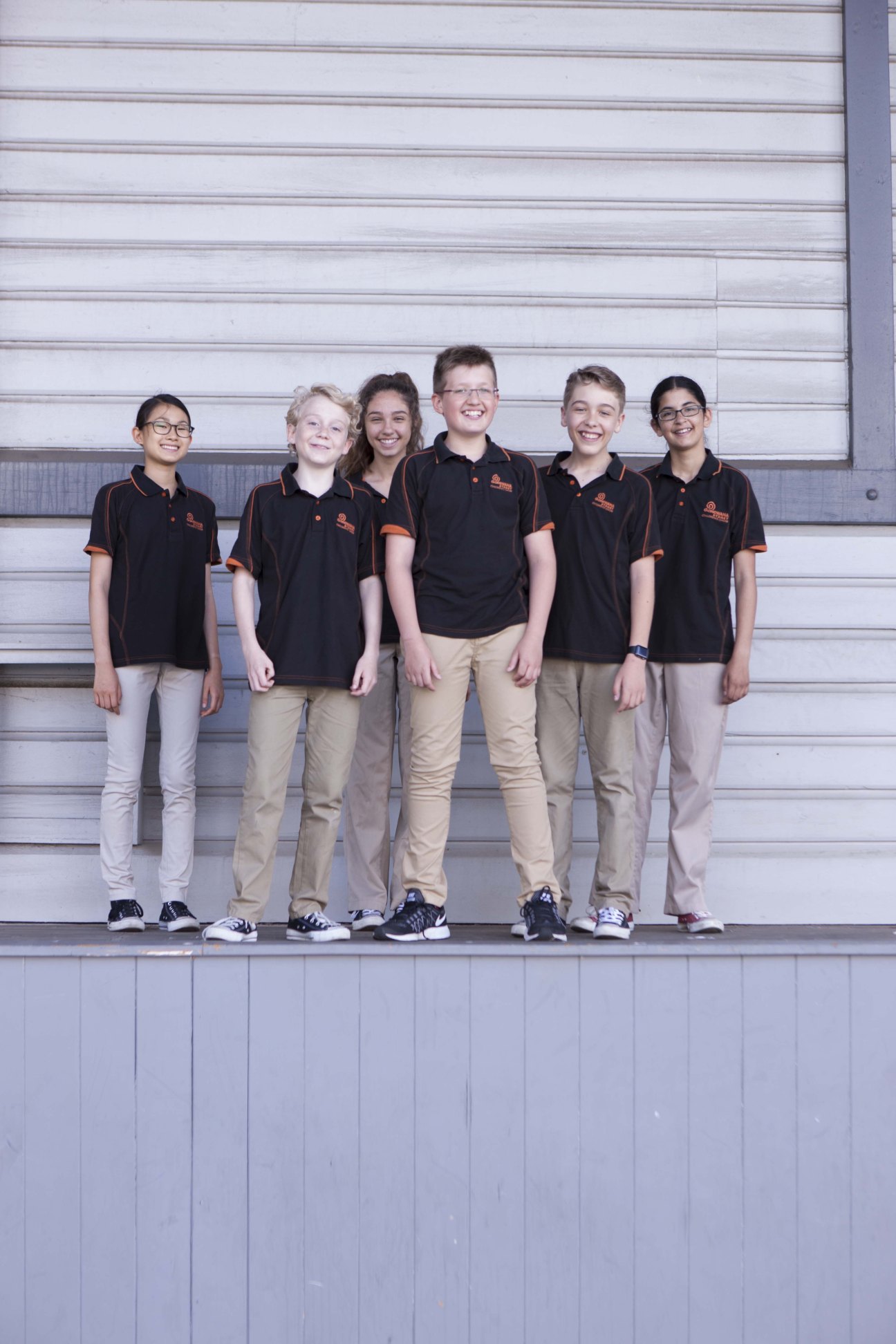 Six children in black and orange choir uniforms stand smiling against a white corrugated metal wall.