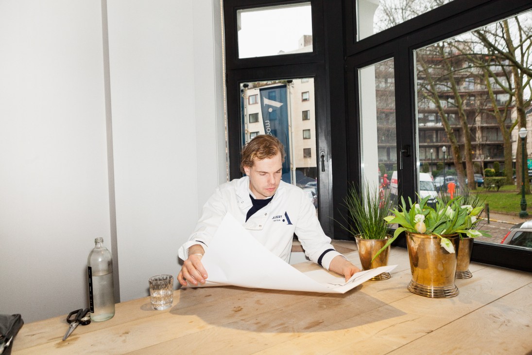 Chef Arno Verbeke in white chef's coat sitting at a wooden table reviewing documents by a window.