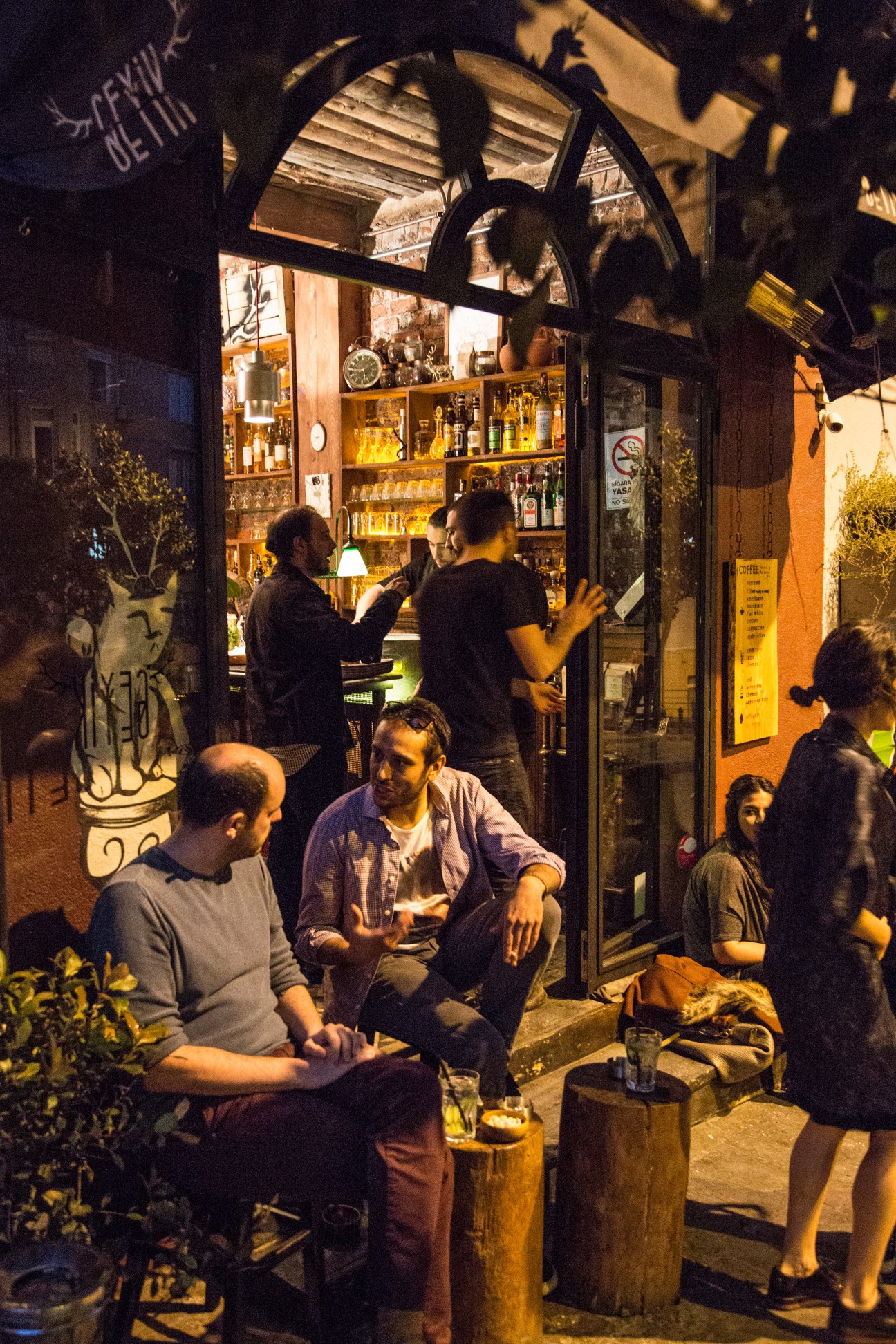 People gathered outside Geyik cafe at night, with illuminated storefront and bottles visible through windows.