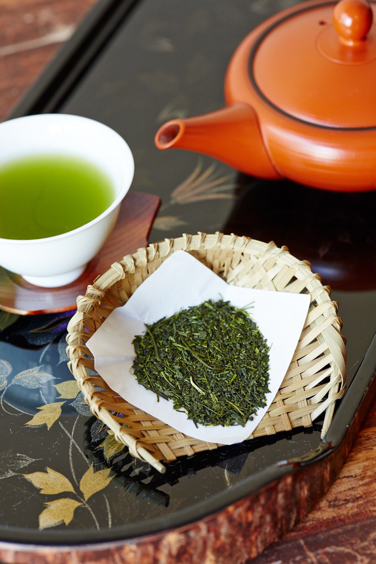 Green tea leaves in a woven basket with a white cup of brewed tea and orange ceramic teapot.