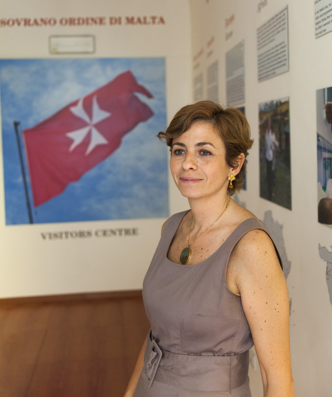 Woman with short brown hair wearing a gray sleeveless dress standing in front of a display with the red Maltese cross flag and text reading "Sovrano Ordine di Malta" and "Visitors Centre"