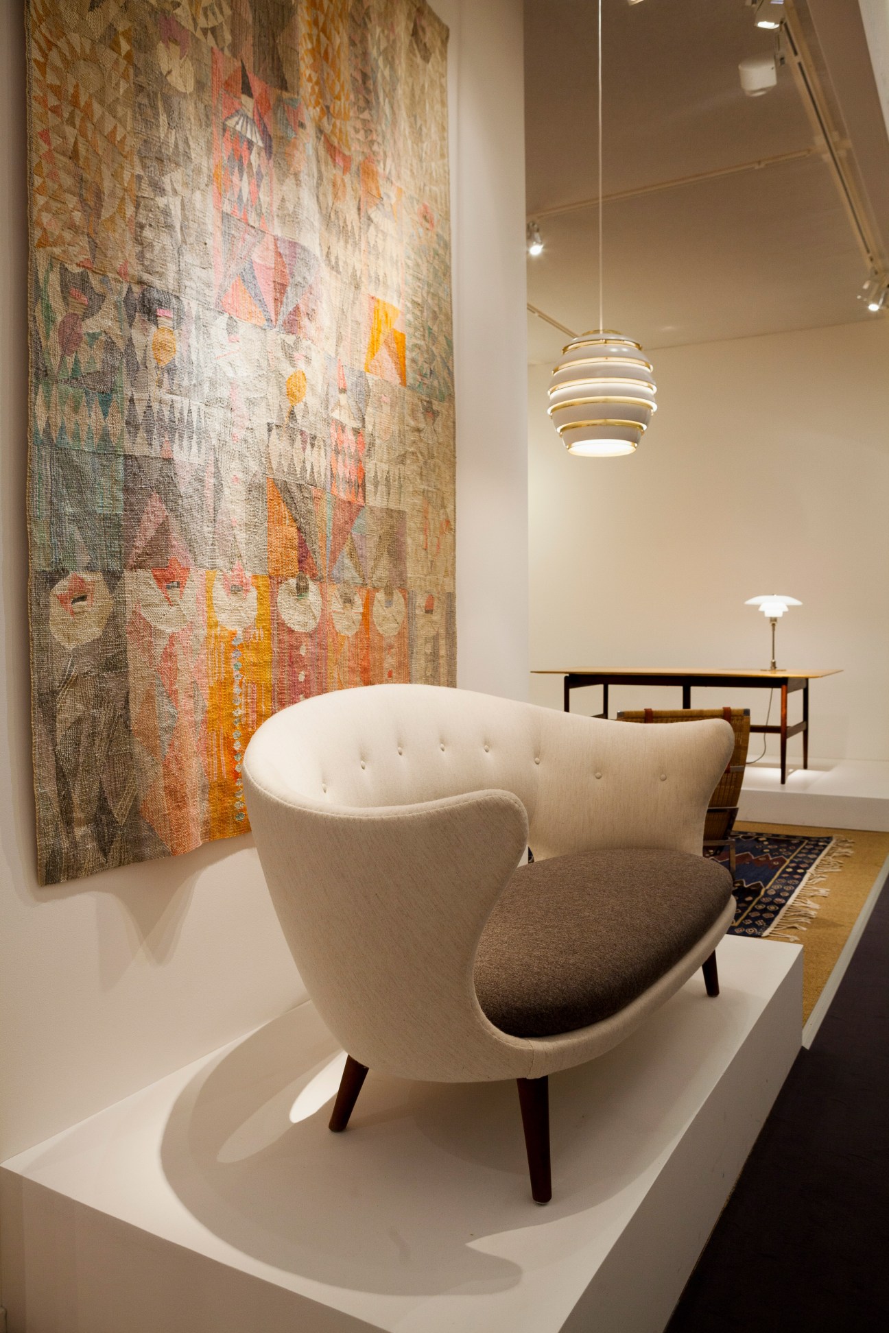 Modernist cream upholstered chair with wooden legs displayed in gallery setting beside colorful textile artwork.