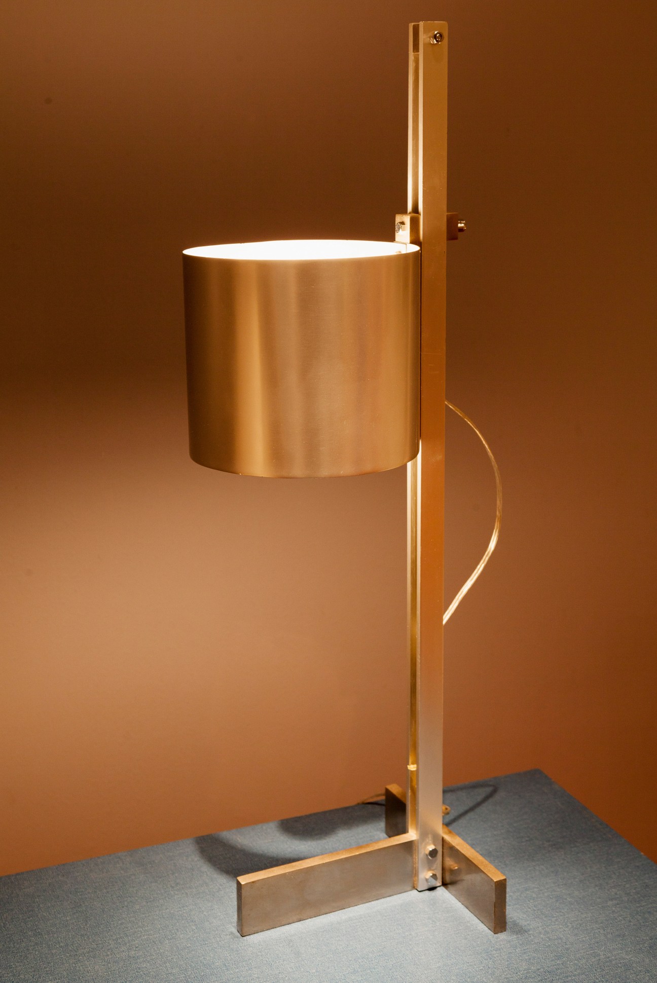 Modernist brass table lamp with cylindrical shade on wooden stand against brown backdrop.