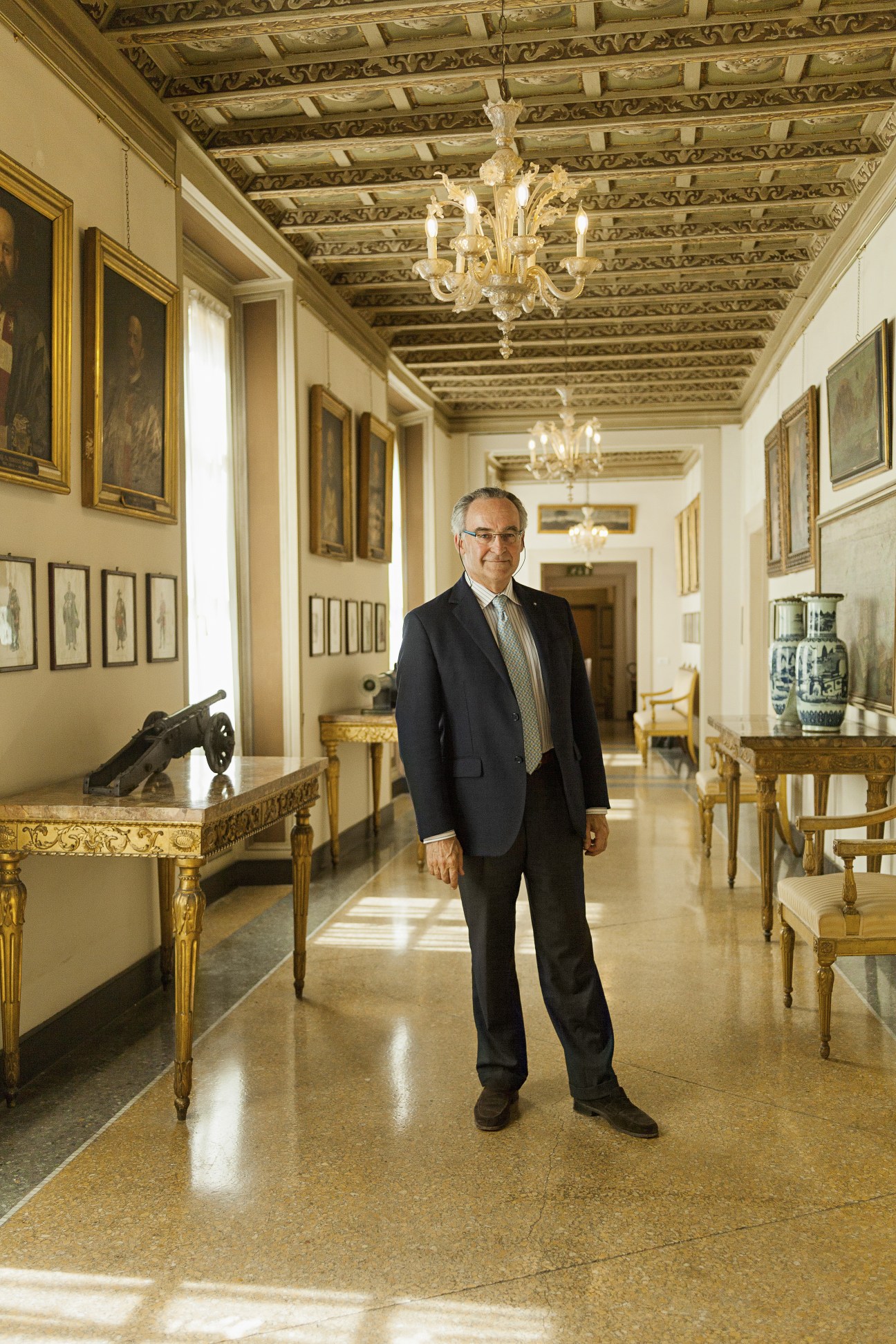 Man in dark suit standing in ornate palace corridor with gilded ceiling and chandeliers