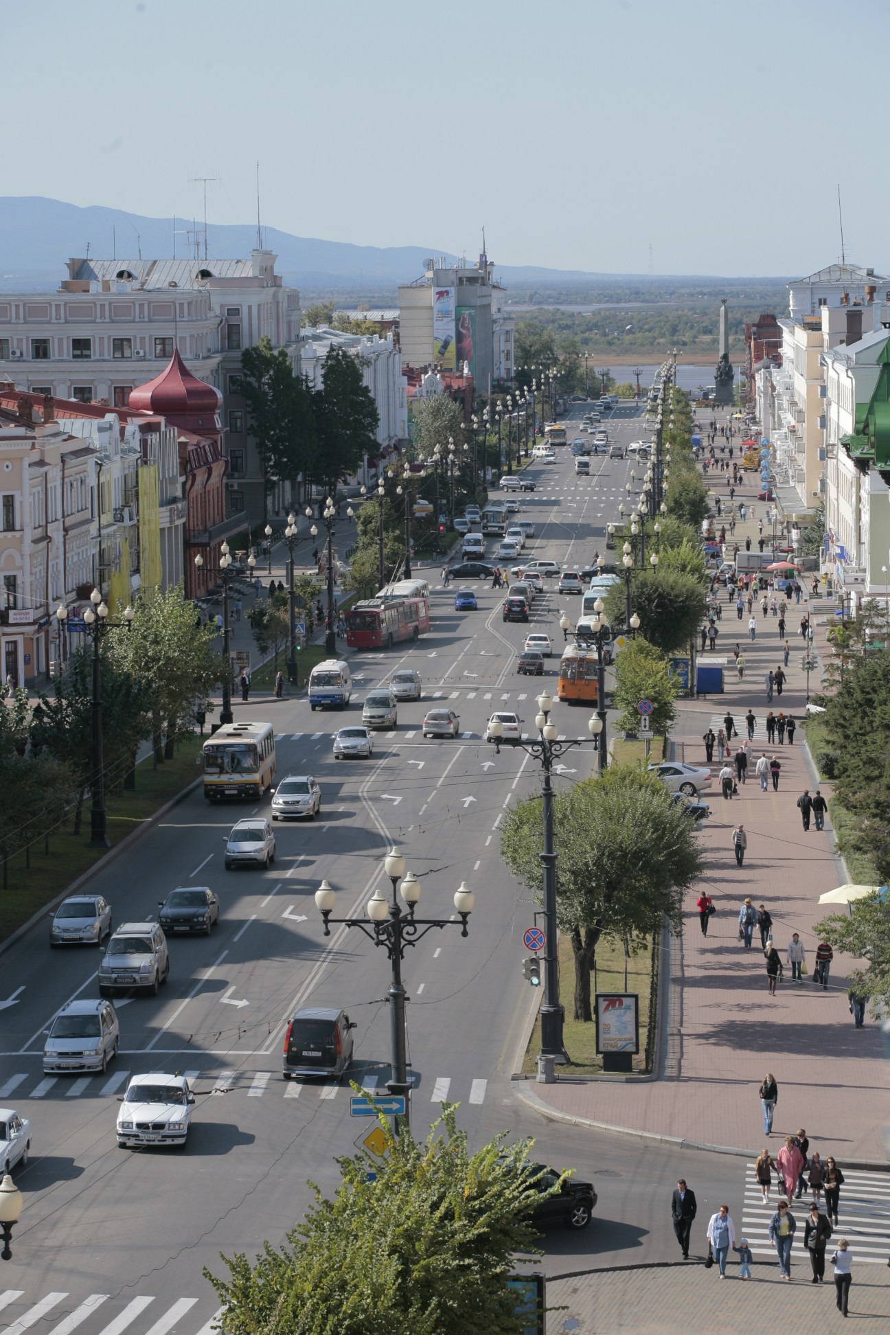 Aerial view of a wide Soviet-era boulevard in Khabarovsk with buildings, traffic, pedestrians, and mountains in the distance.
