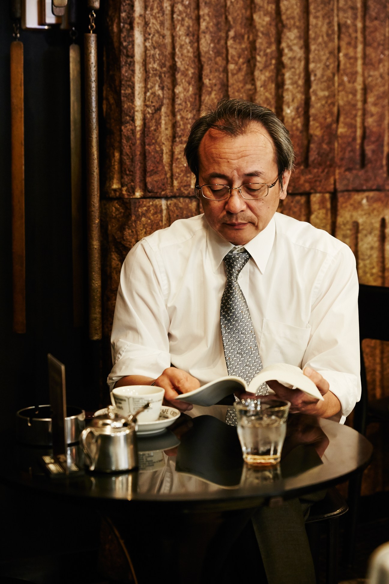 A man in business attire sits at a café table reading, with a coffee cup and glass of water beside him.