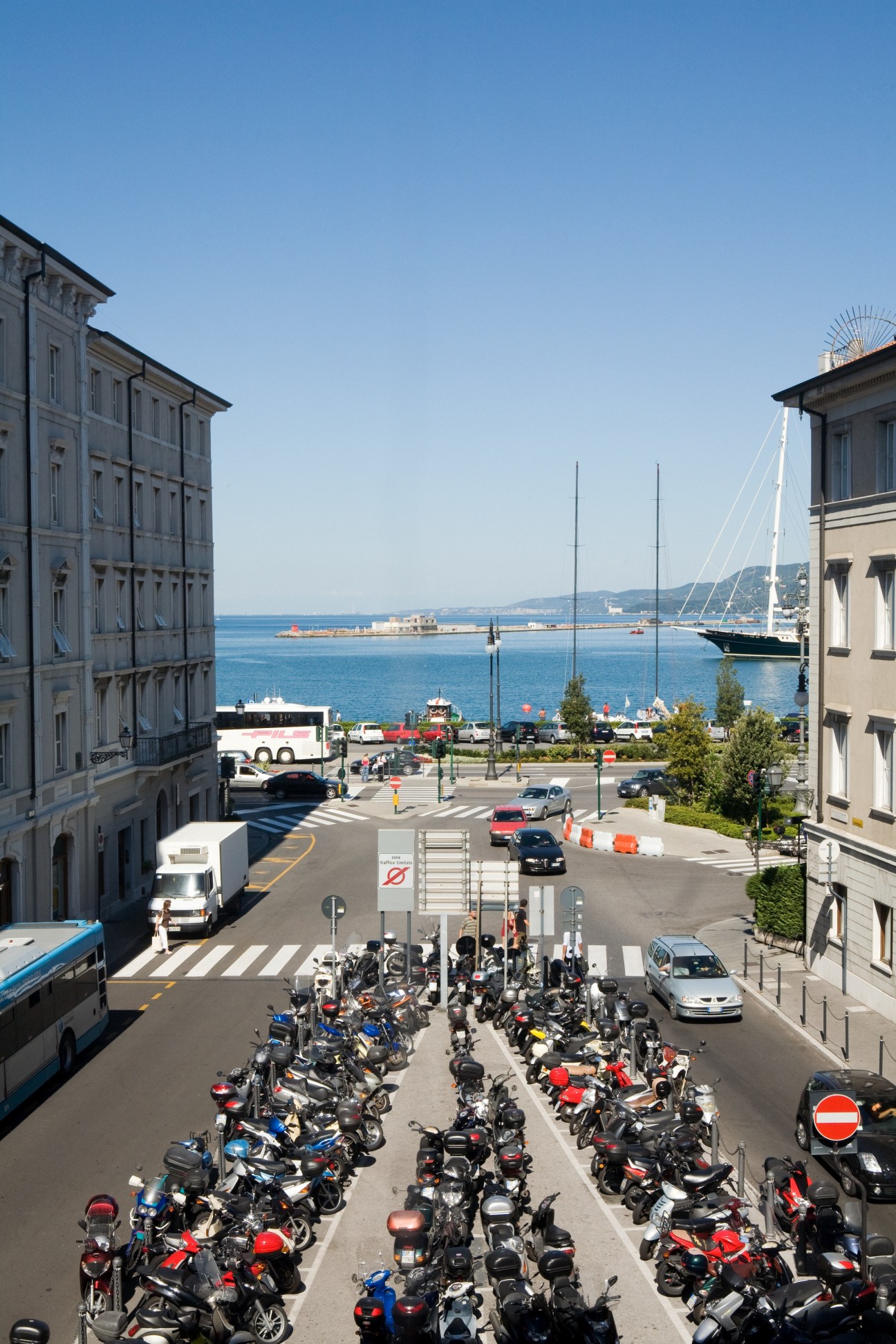 Aerial view of Trieste waterfront showing parked scooters on a street with historic buildings and sailboats in the harbor.