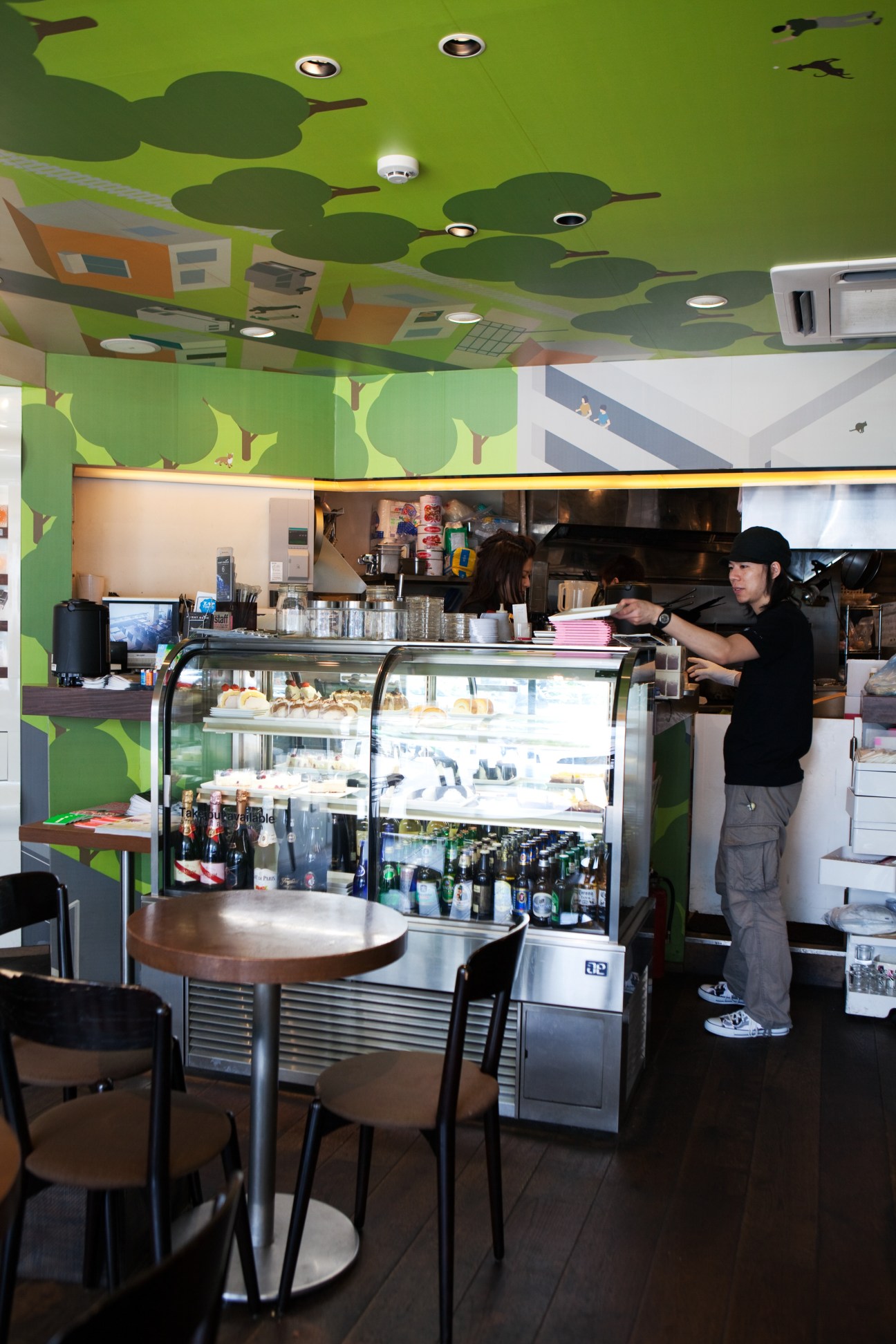 Interior of Sign Cafe Tokyo showing bright green ceiling with tree design, counter with pastries, and staff member behind display case.