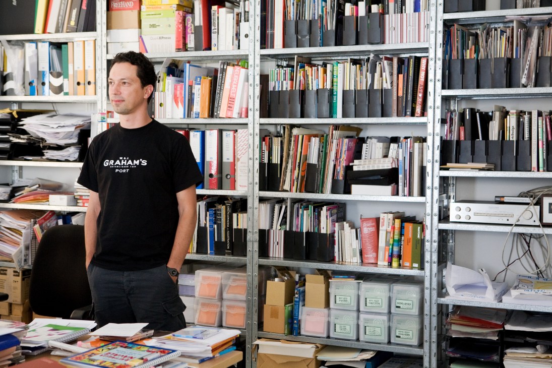 Graphic designer Rado Jagodic stands in his studio surrounded by shelves of books, design materials, and work samples.