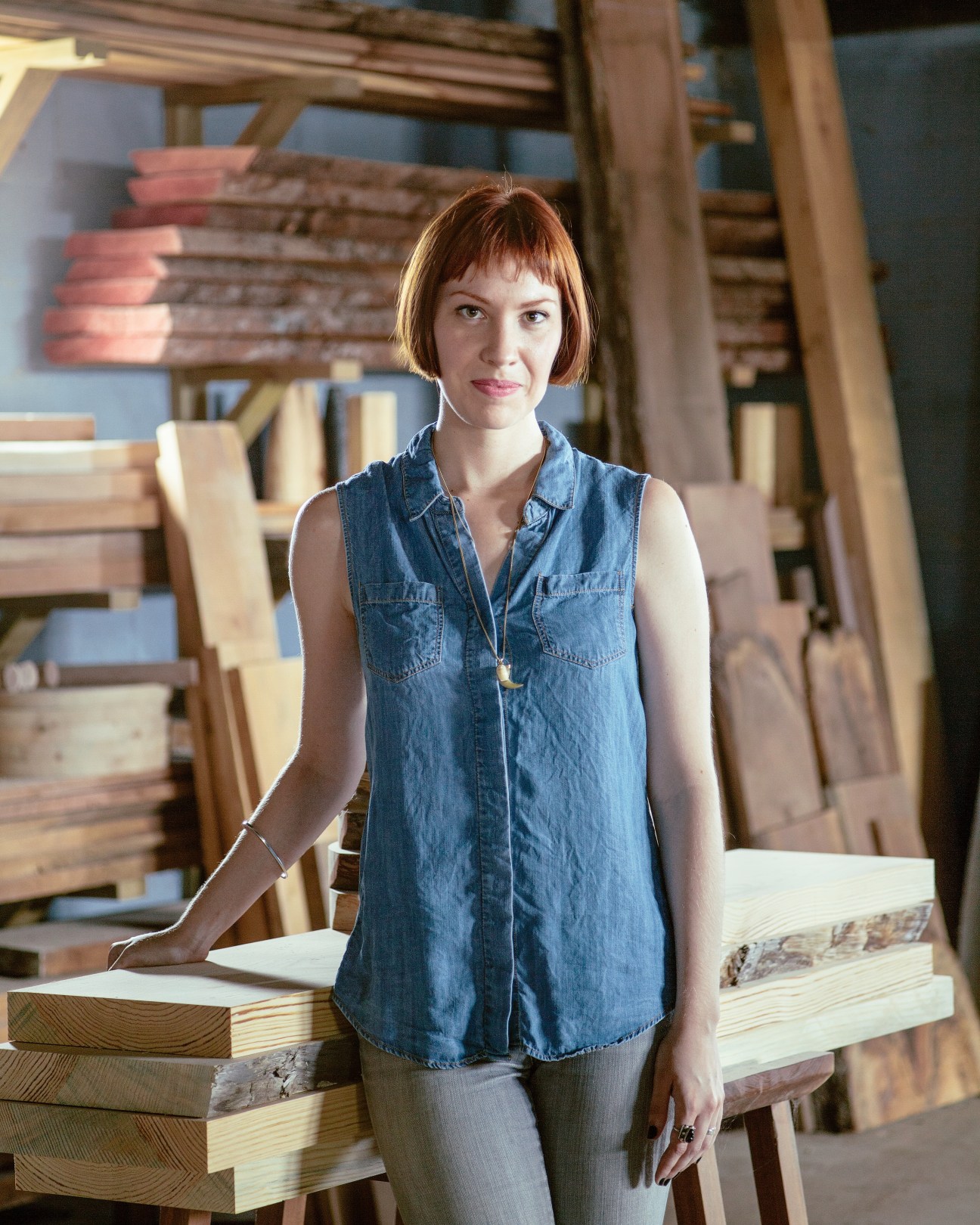 Celia Gibson stands in a furniture workshop surrounded by wooden pieces and timber materials.