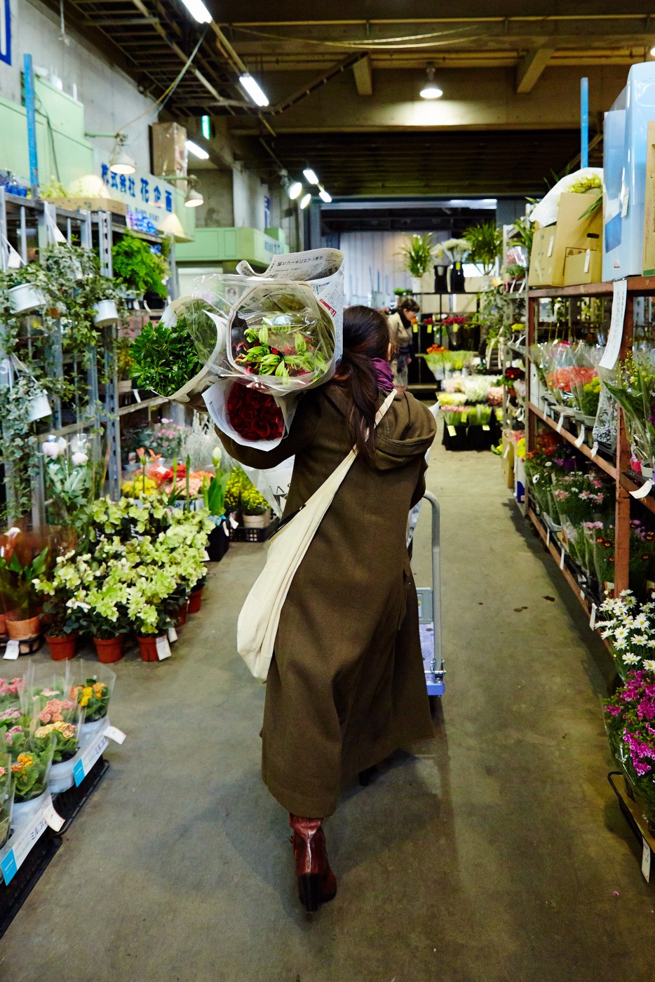A buyer carries wrapped flower bundles through an aisle lined with potted plants and flowers at Ota market.