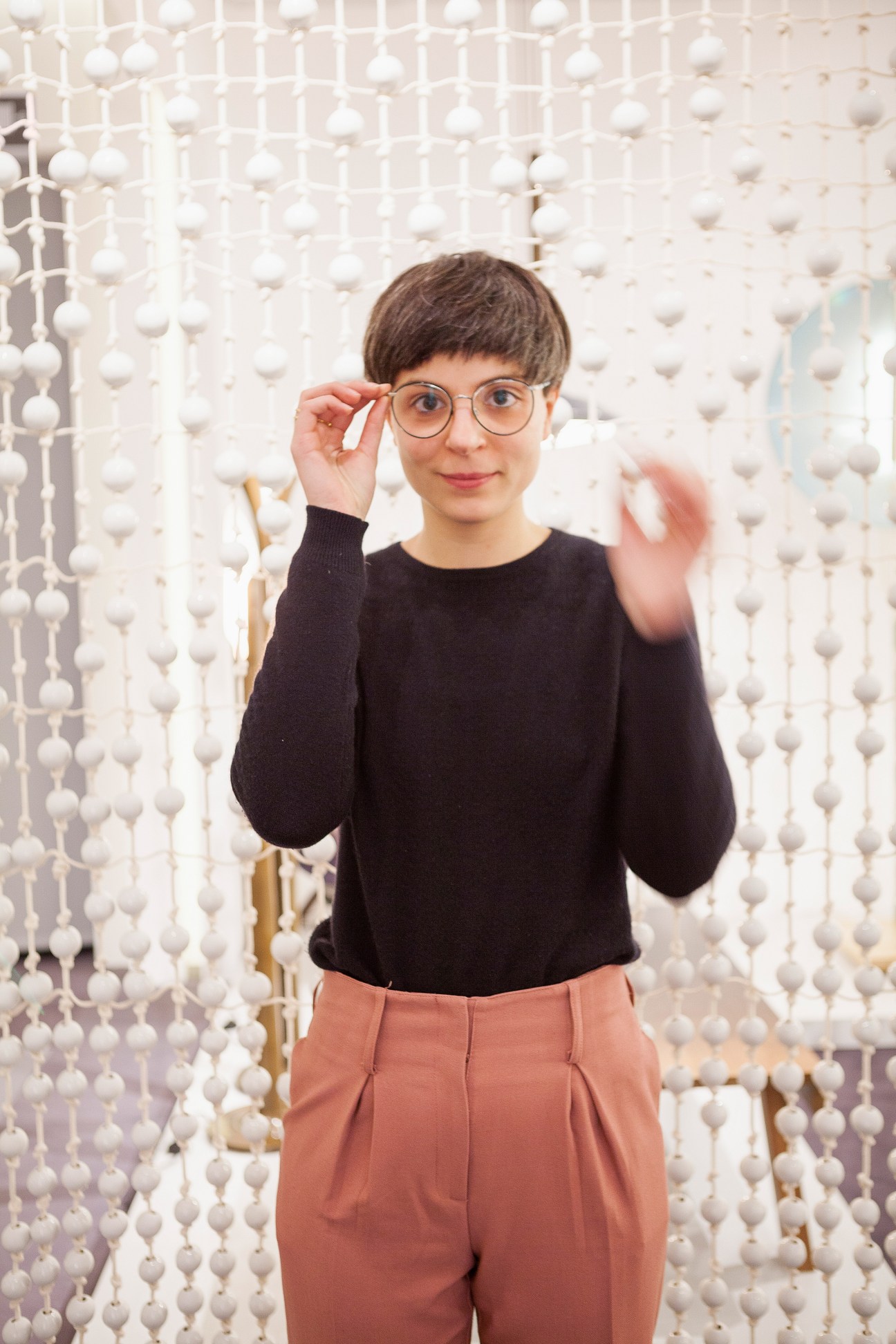 Young person wearing round glasses and black sweater in front of white beaded curtain backdrop at Galerie Kreo.