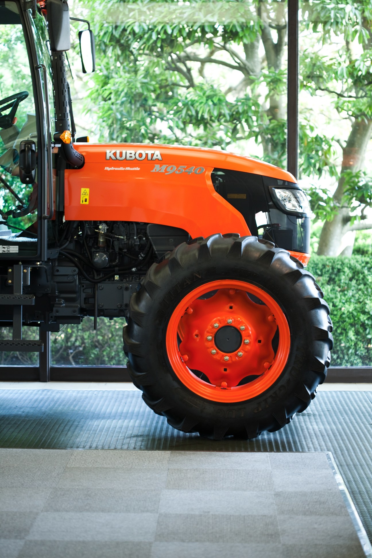 Orange Kubota M0540 tractor with large black tires displayed indoors with green foliage visible in background.