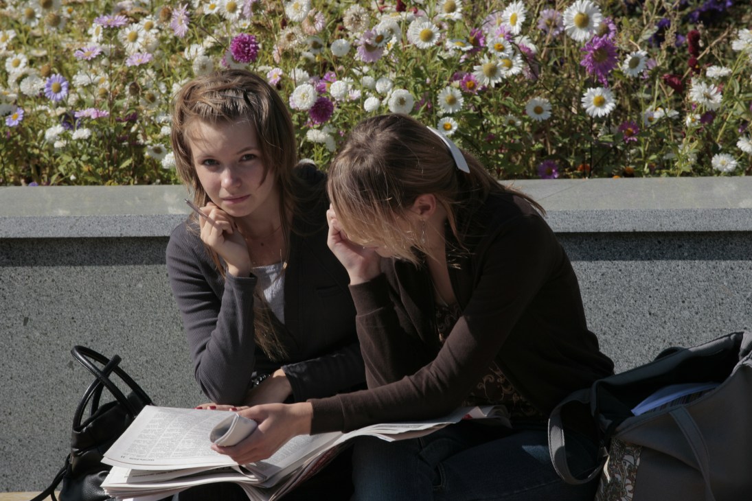 Two young female students study together on a bench with flowering plants in the background.