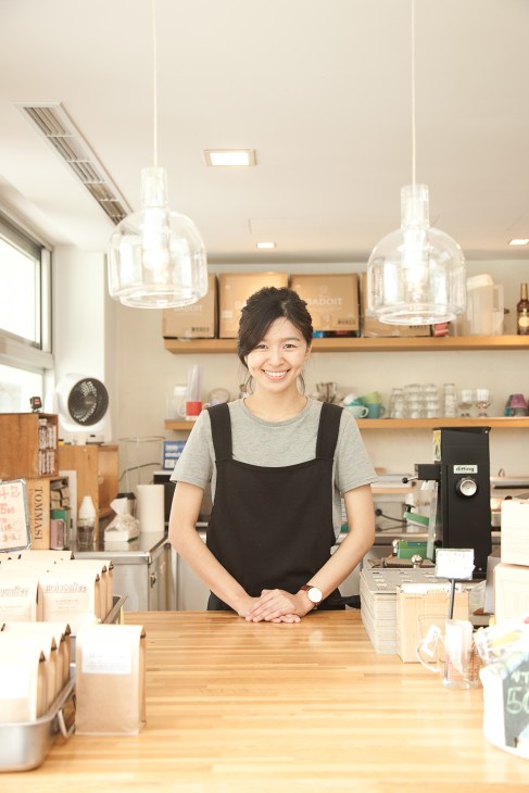 A smiling barista stands behind a wooden counter in a bright, minimalist coffee shop with pendant lights and shelving.