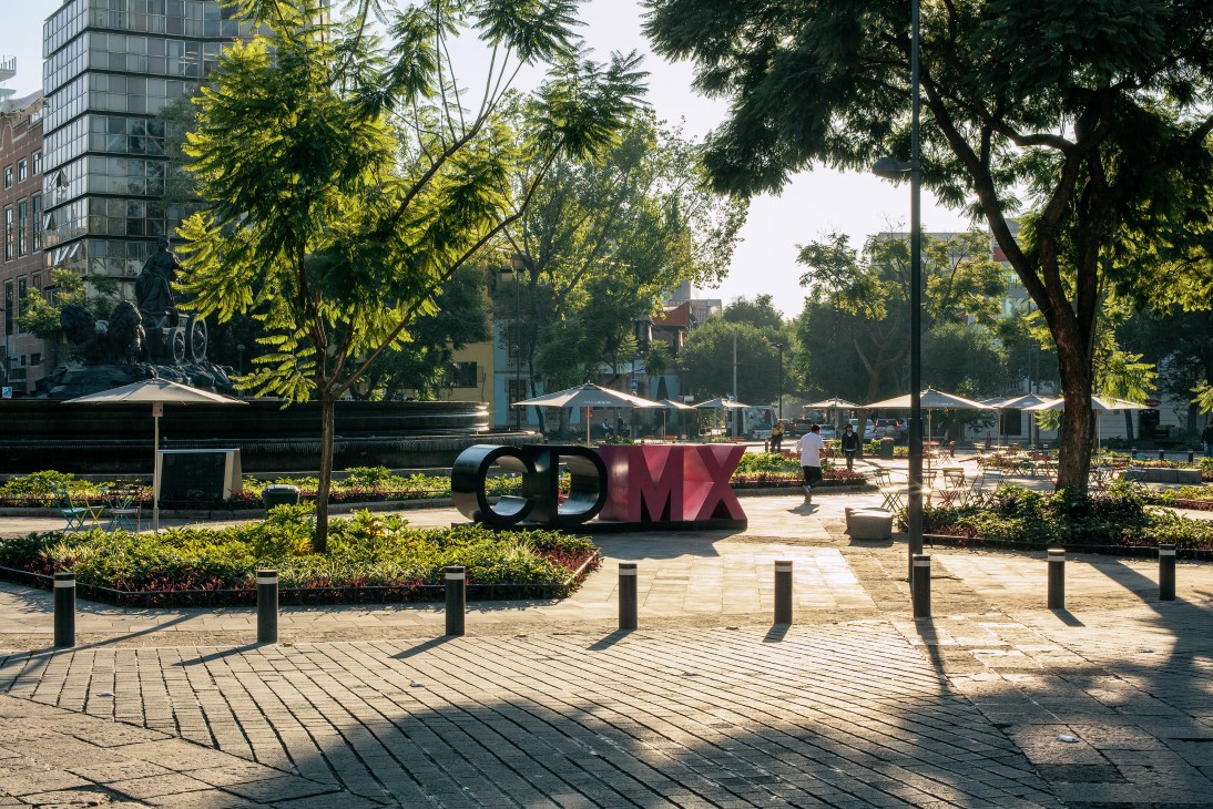 Tree-lined public plaza in Roma Norte with seating areas, umbrellas, and a large CDMX sculpture.