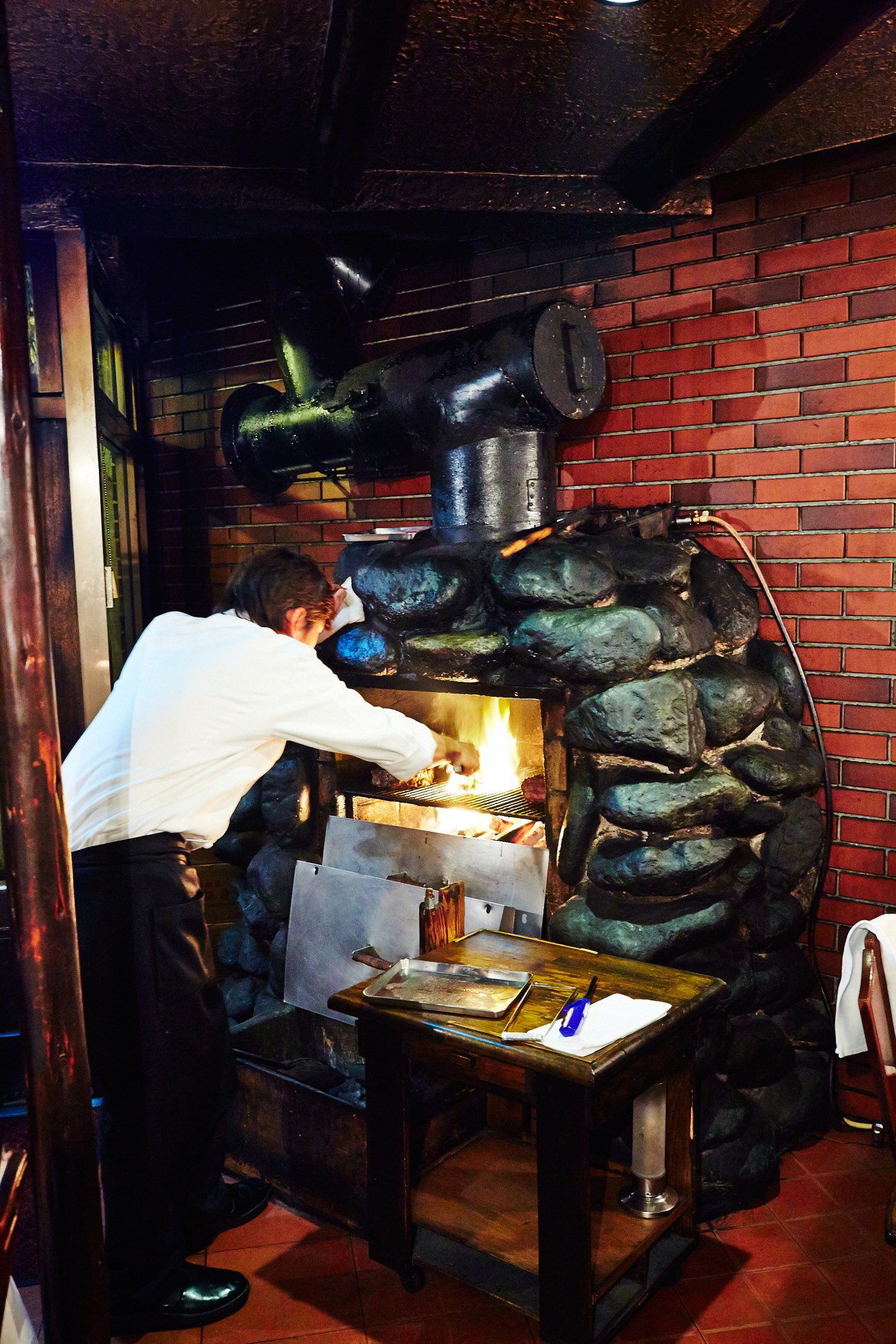 Chef cooking steaks on a charcoal grill in a brick-walled kitchen.