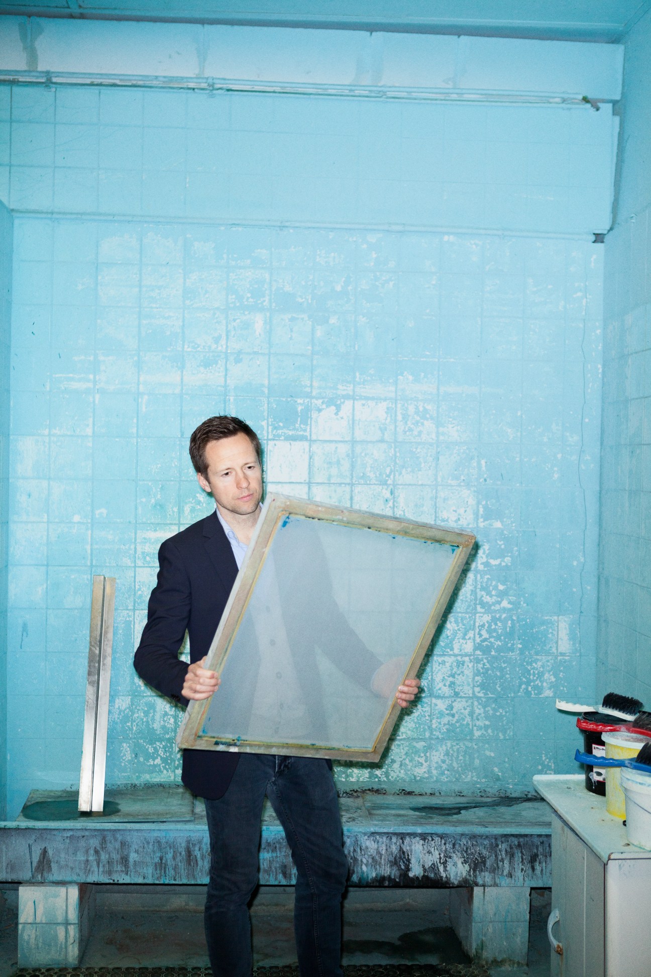 Man in dark blazer holds framed artwork against turquoise tiled wall in studio setting