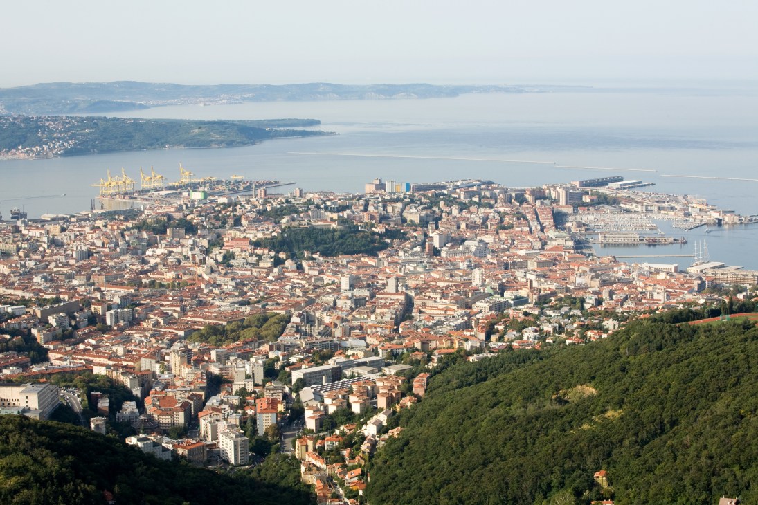 Aerial view of Trieste city with commercial port visible at top left and Adriatic Sea beyond.