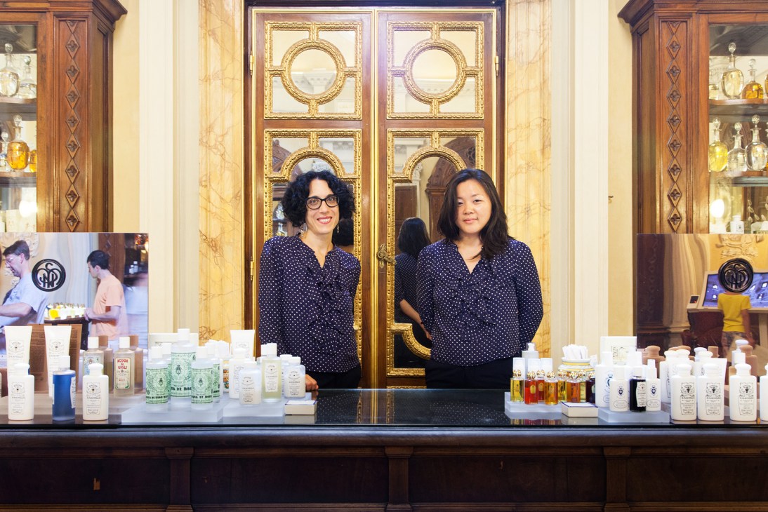 Two women in navy polka dot shirts stand behind a counter at Officina di Santa Maria Novella in Florence, with ornate mirrors and product displays.