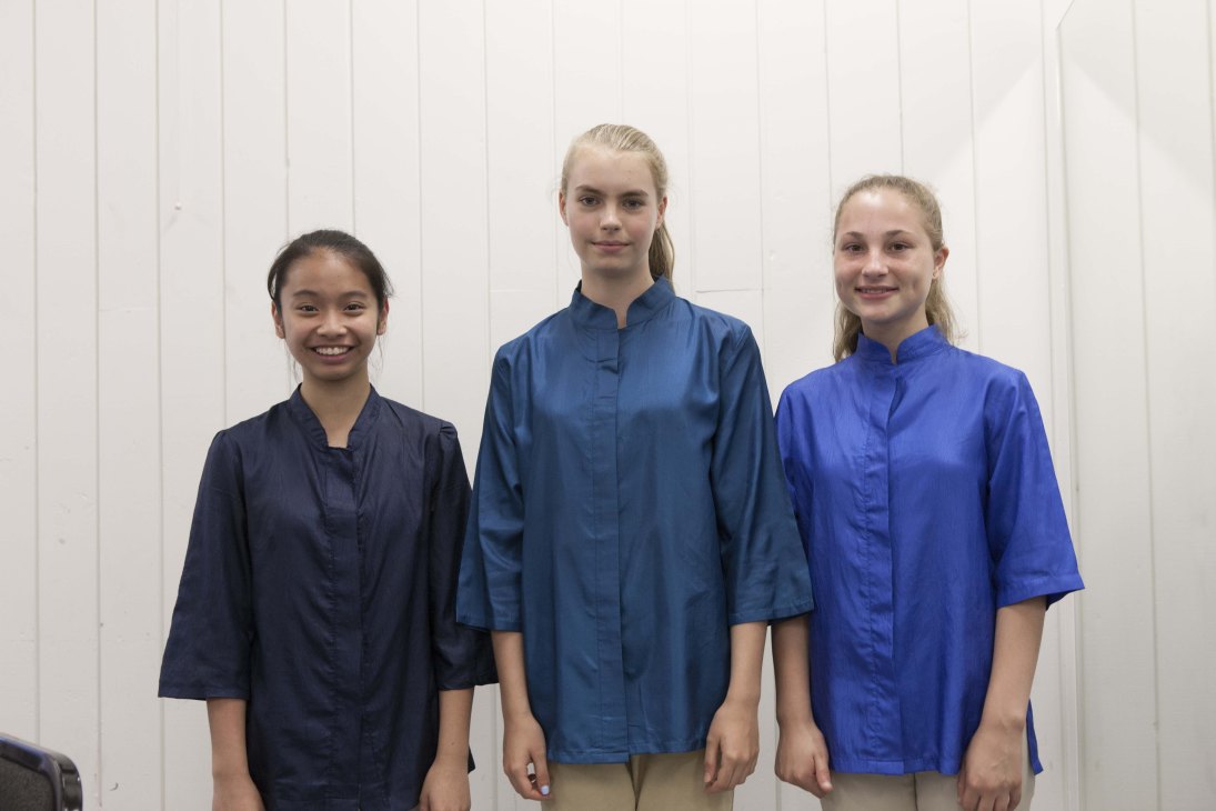Three young choir members in blue uniform shirts stand together against a white wall during costume fitting.