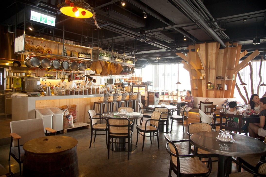 Modern restaurant interior with wooden accents, hanging cookware, and diners seated at tables.