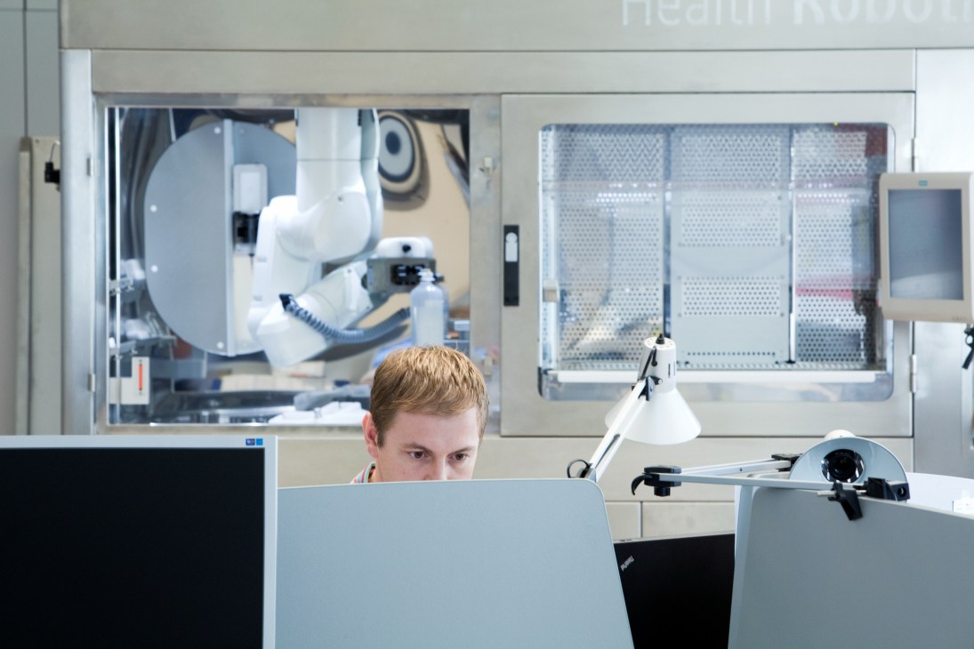 Researcher working at a desk in a robotics laboratory with robotic equipment visible through glass panels behind him.