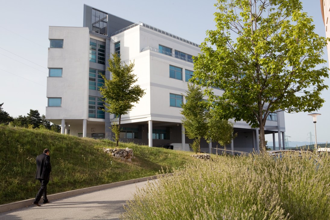 Modern white building with glass windows at Area Science Park housing start-ups, surrounded by landscaped grounds with vegetation.