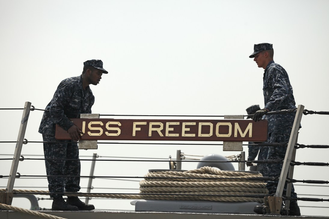 Two US Navy personnel in digital camouflage uniforms stand on either side of the USS Freedom nameplate on the ship's railing.