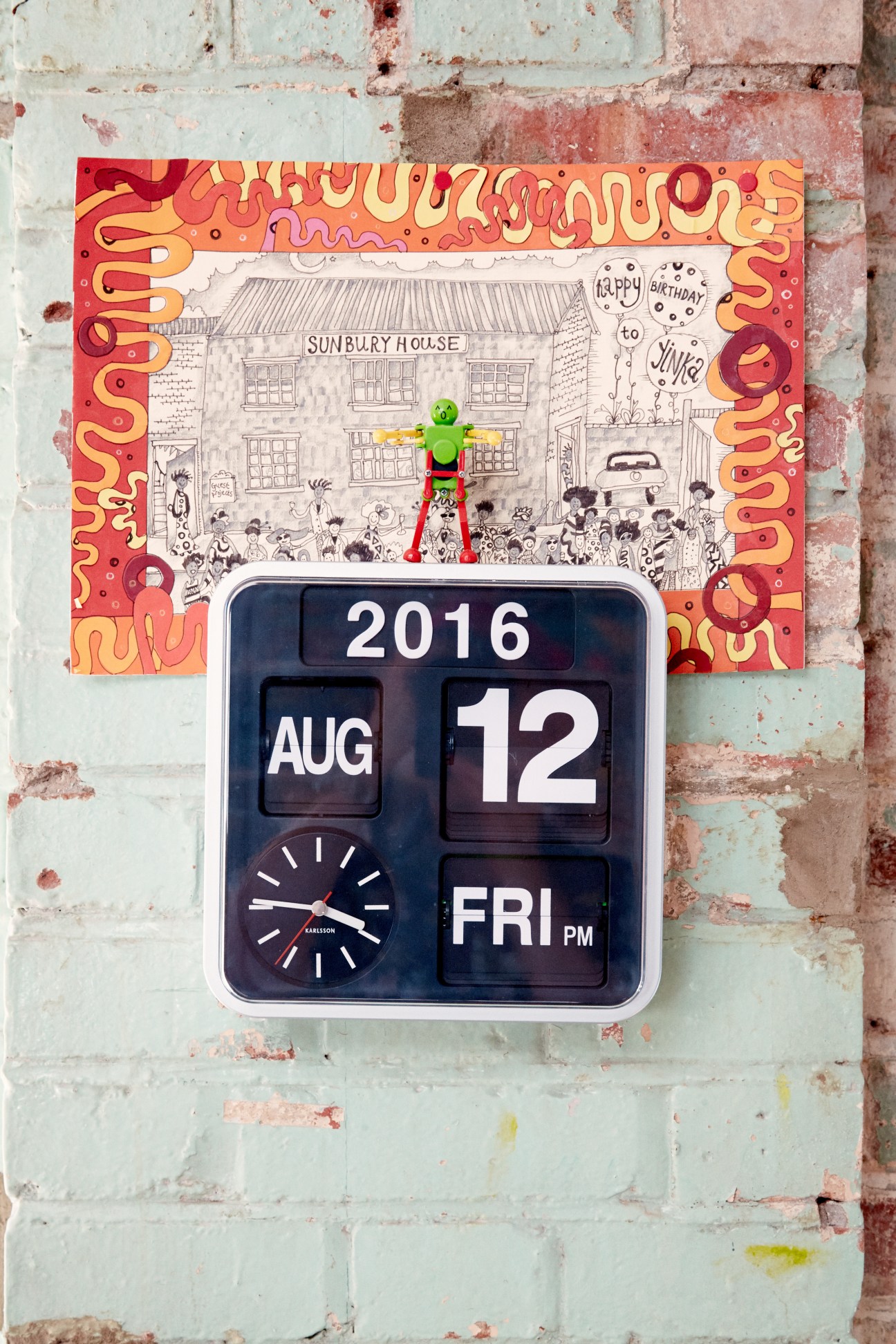 A flip clock displaying August 12, 2016 mounted on a brick wall below a colorful illustration featuring Sunbury House with vibrant red and orange patterned border.